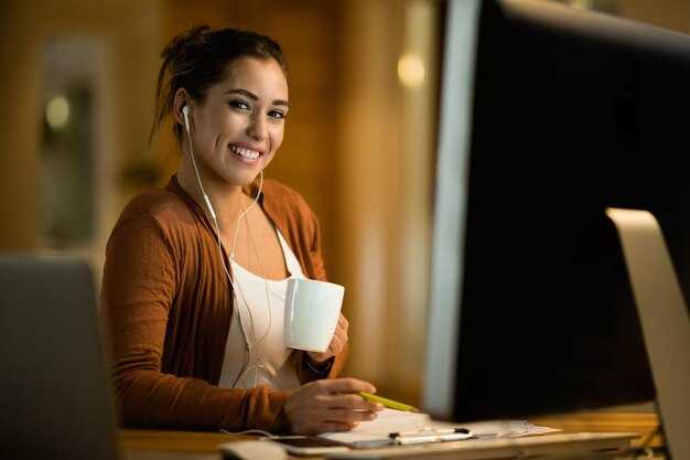 Young happy woman drinking coffee while studying late at night and looking at camera