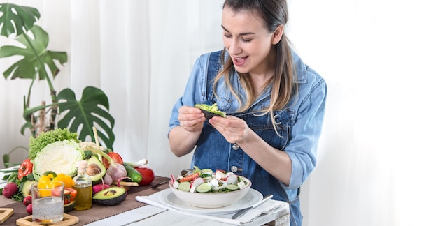 Young and happy woman eating salad with organic vegetables at the table on a light background , in denim clothes. The concept of a healthy home-made food.
