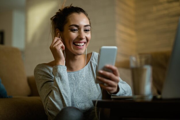 Молодая счастливая женщина развлекается, используя мобильный телефон и читая текстовое сообщение в своей гостиной