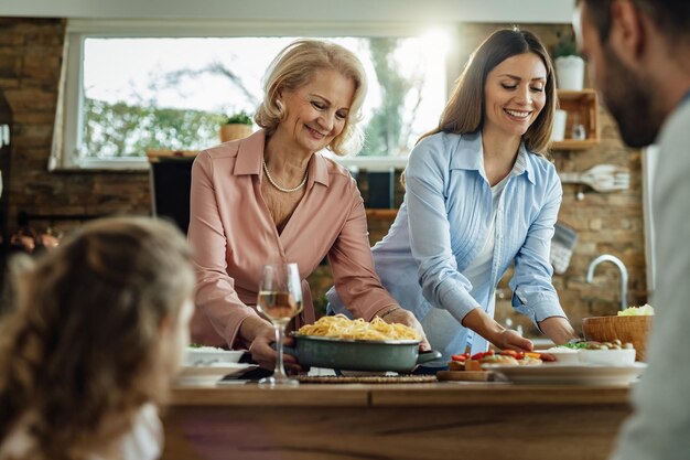若い幸せな女性と彼女の母親は、家で家族の昼食をとりながら、テーブルに食べ物を持ってきます
