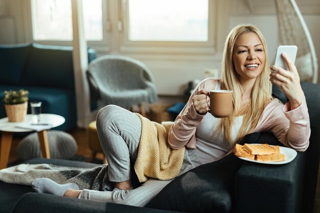 커피를 마시고 소파에서 휴식을 취하는 동안 휴대폰으로 문자 메시지를 읽는 젊은 행복한 여성.