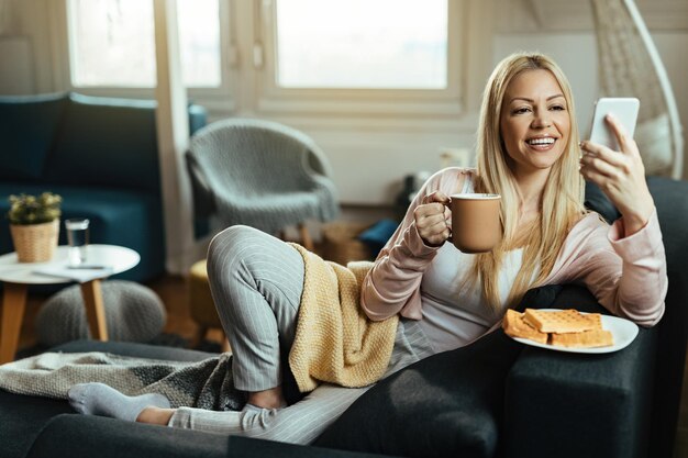 コーヒーを飲みながらソファでリラックスしながら携帯電話でテキストメッセージを読んで若い幸せな女性。