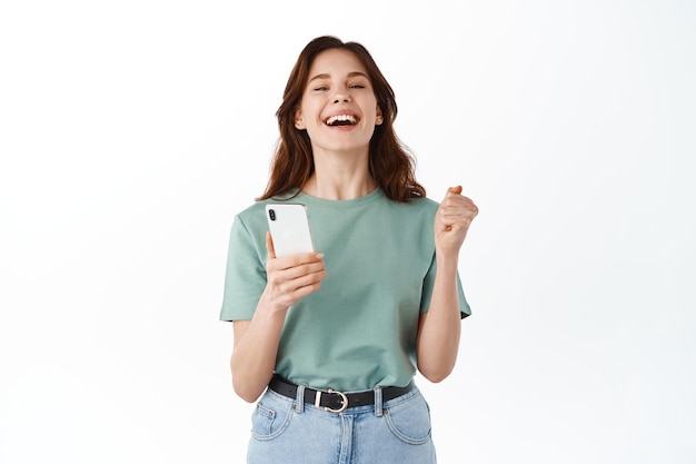 Young happy woman say yes, winning prize or daily goal on mobile app, holding smartphone and rejoicing, celebrating victory in application, standing against white background