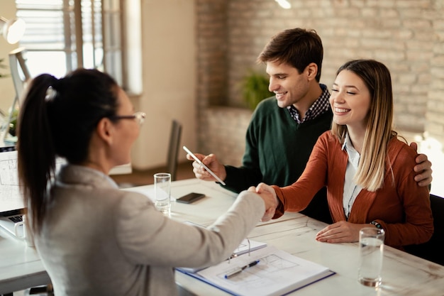 Young happy woman shaking hands with real estate agent while her husband is using touchpad in the office