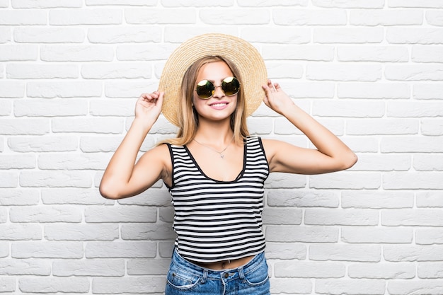 흰색 벽돌 벽 위에 선글라스와 여름 모자에 웃 고 젊은 행복 한 여자