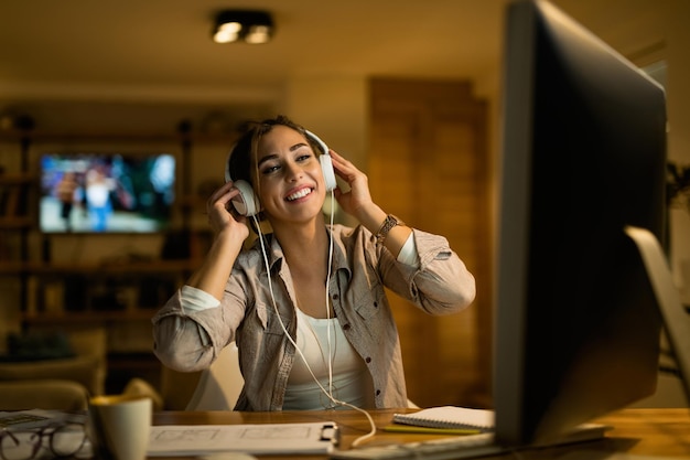 Молодая счастливая женщина сидит в сети на компьютере, слушая музыку в наушниках вечером дома