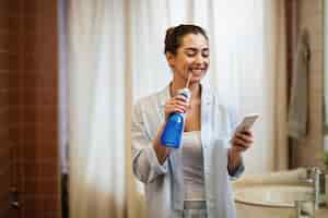 Free photo young happy woman using dental water flosser and cleaning her teeth while texting on mobile phone in the bathroom