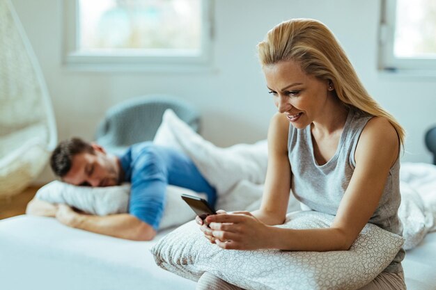 Young happy woman using mobile phone while sitting on the bed. Her boyfriends is sleeping behind her.