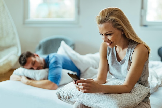 ベッドに座って携帯電話を使用して若い幸せな女性。彼女のボーイフレンドは彼女の後ろで寝ています。