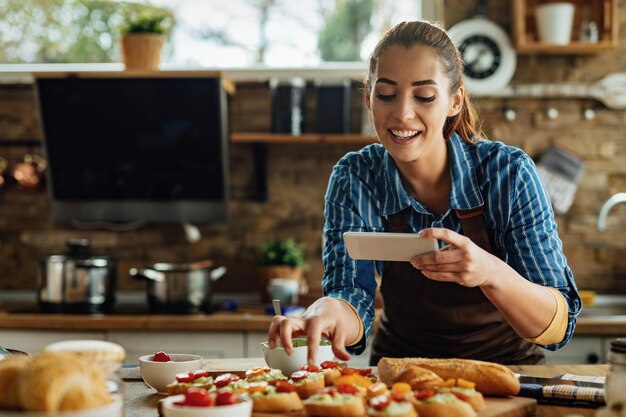 スマートフォンを使用して、キッチンで準備した食べ物を撮影する若い幸せな女性