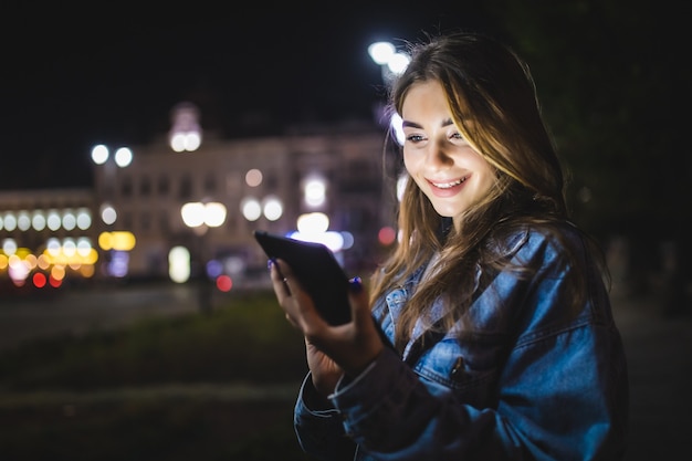 Молодая счастливая женщина с помощью планшета на открытом воздухе над размытыми огнями ночного города