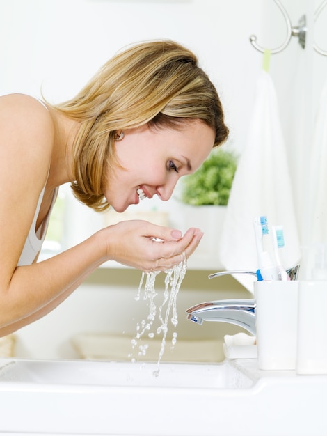 Foto gratuita giovane donna felice lavaggio viso con acqua in piedi in bagno