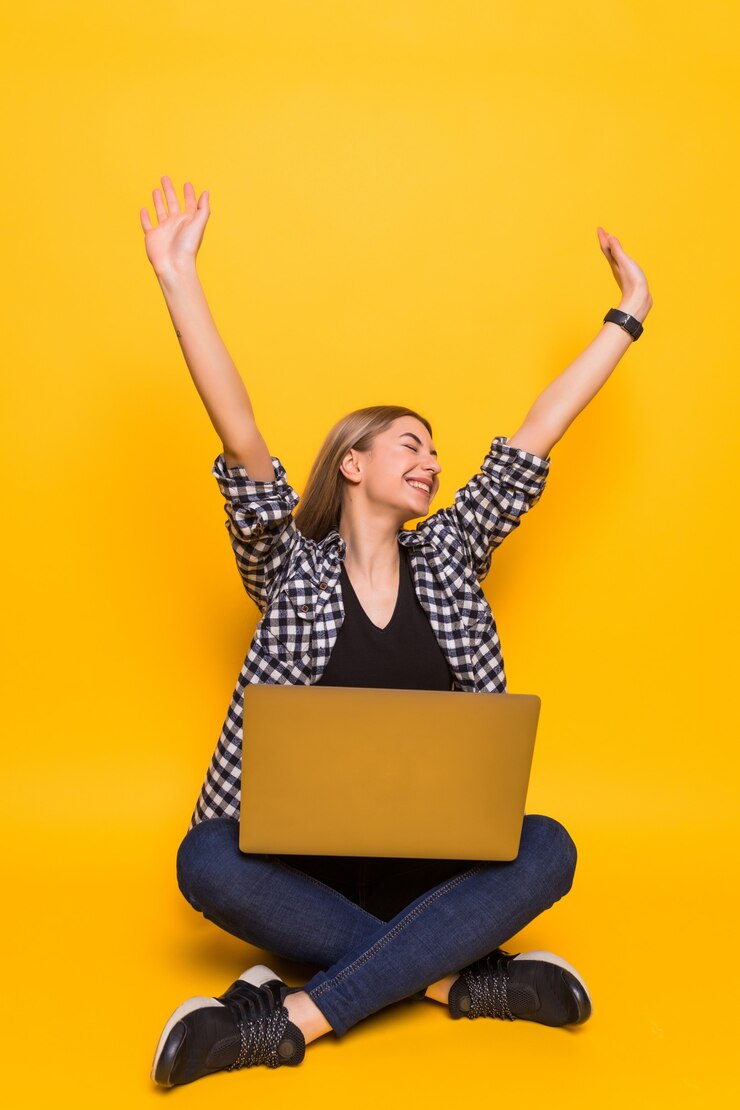 मुफ़्त फोटो young happy woman in white t-shirt is sitting using laptop and celebrating victory and success over yellow wall