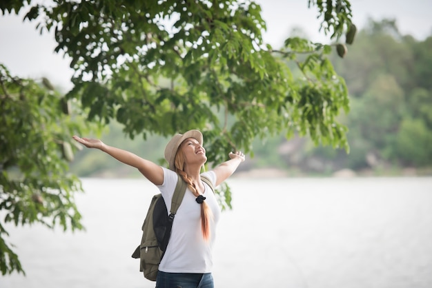 Молодая счастливая женщина с рюкзак, стоя с поднятыми руками и глядя на реку.