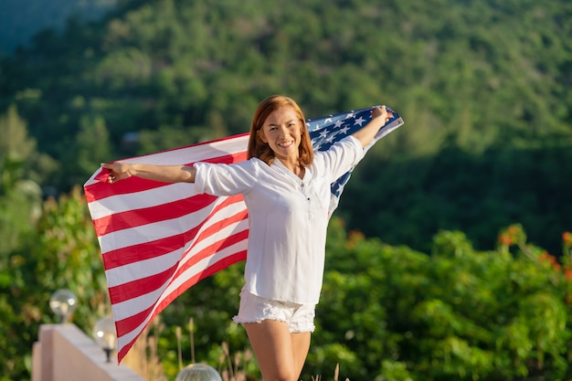 Молодая счастливая женщина с флагом Соединенных Штатов, наслаждаясь закатом на природе.