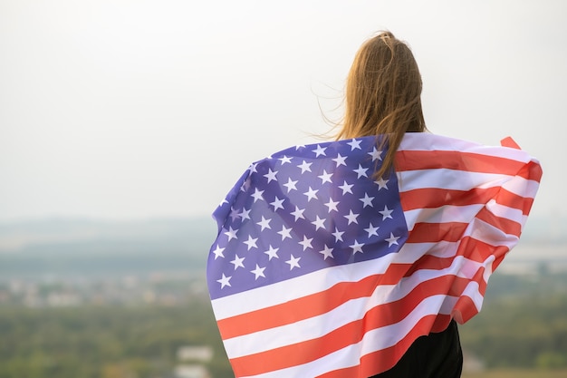Young happy woman with long hair holding waving on wind american national flag on her sholders resting outdoors enjoying warm summer day.