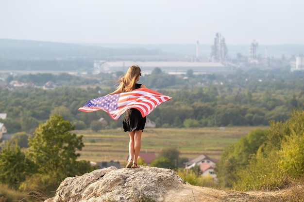 Young happy woman with long hair holding waving on wind american national flag on her sholders resting outdoors enjoying warm summer day.
