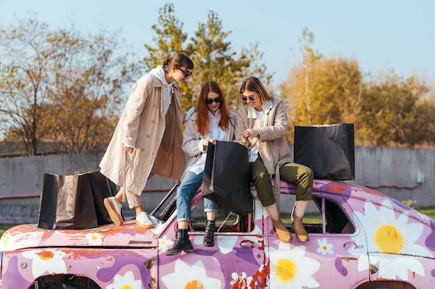 Free photo young happy women with shopping bags posing near an old decorated car