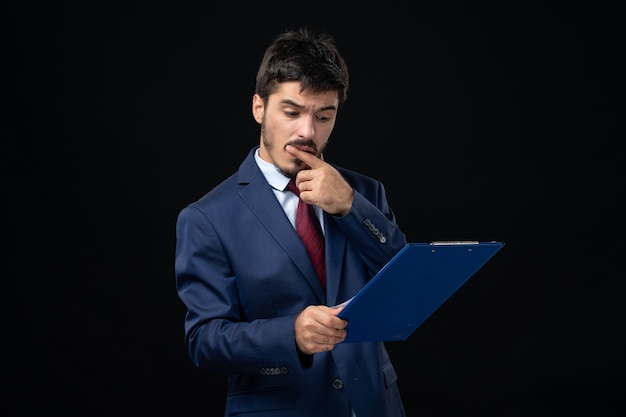 Free photo young hardworking male office worker in suit holding documents and checking statistics in it on isolated dark wall