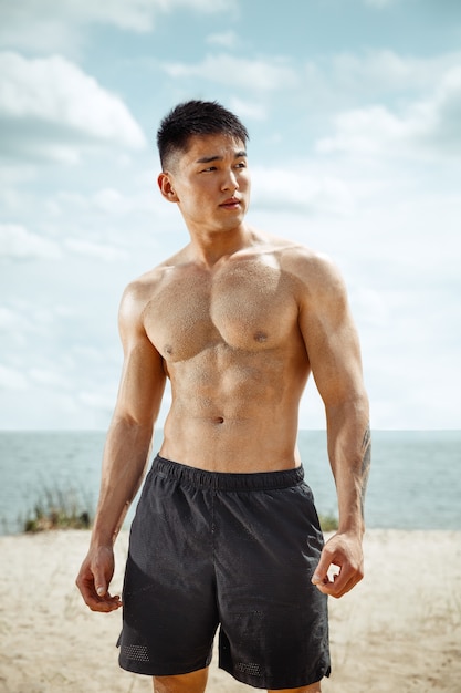 ビーチで運動をしている若い健康な男性アスリート。晴れた日の川沿いでのサイン男性モデル上半身裸のトレーニング空気。健康的なライフスタイル、スポーツ、フィットネス、ボディービルの概念。
