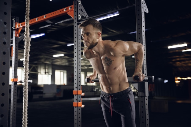 Young healthy man, athlete doing exercises, pull-ups in gym. Single male model practicing hard and training his upper body. Concept of healthy lifestyle, sport, fitness, bodybuilding, wellbeing.