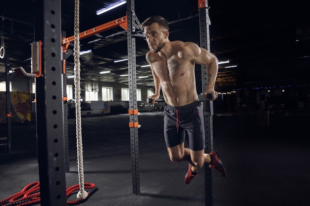 Young healthy man, athlete doing exercises, pull-ups in gym. Single male model practicing hard and training his upper body. Concept of healthy lifestyle, sport, fitness, bodybuilding, wellbeing.
