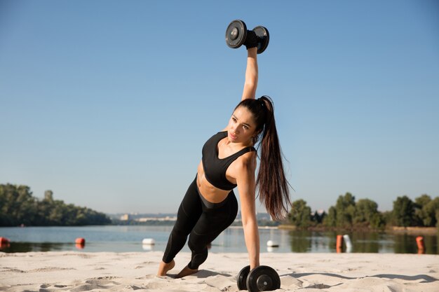Молодая здоровая женщина тренирует верхнюю часть тела с весами на пляже