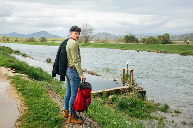 Young hiker standing near flowing river with holding backpack looking at camera