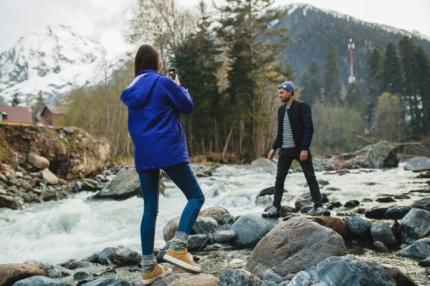 Young hipster beautiful couple in love holding smartphone, taking pictures, at river in winter forest