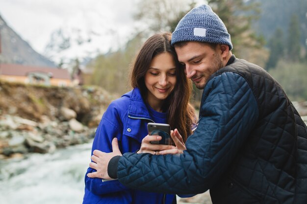 Young hipster beautiful couple in love holding smartphone, taking pictures, at river in winter forest