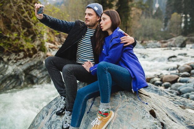 スマートフォンを押し、写真を撮って、冬の森の川の岩の上に座って恋に流行に敏感な若い美しいカップル