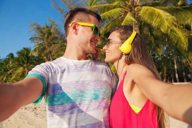 Young hipster beautiful couple in love making selfie photo on tropical beach, summer vacation, happy together, honeymoon, colorful style, sunglasses, headphones, smiling, happy, having fun, positive