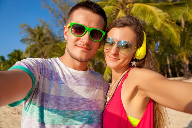 Молодая хипстерская красивая влюбленная пара, делающая селфи-фото на тропическом пляже, летние каникулы, счастливы вместе, медовый месяц, красочный стиль, солнцезащитные очки, наушники, улыбка, счастливая, веселая, позитивная