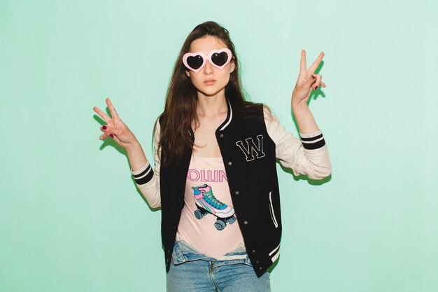 Free photo young hipster beautiful woman, funny heart sunglasses, against blue wall, cool face expression