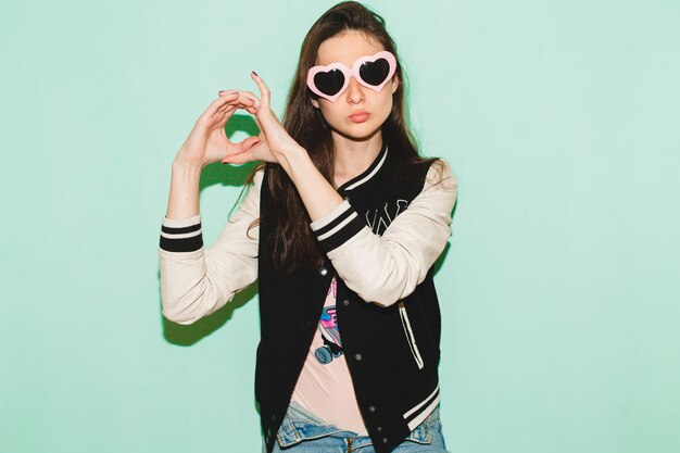 Young hipster beautiful woman, funny heart sunglasses, against blue wall, cool face expression