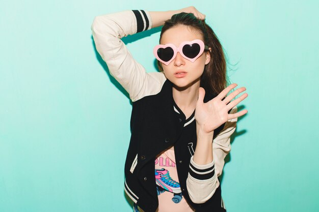 Free photo young hipster beautiful woman, funny heart sunglasses, against blue wall, non-isolated, cool face expression