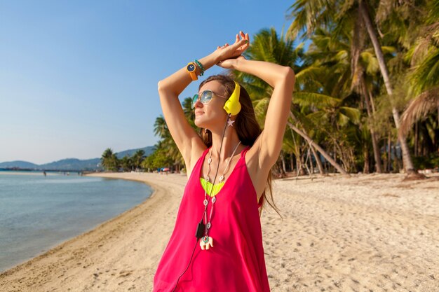 Young hipster beautiful woman, tropical beach, vacation, colorful, summer trend style, sunglasses, headphones, listening to music, palm trees background, smiling happy, fun, details, close up portrait