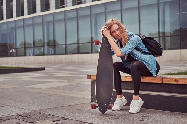 Young hipster blonde girl in casual clothes, sitting on a bench against a skyscraper, resting after riding on a skateboard.