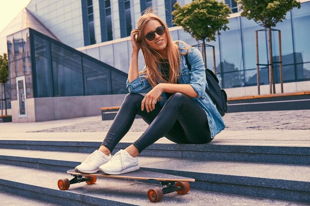 Молодая блондинка-хипстер в повседневной одежде и солнцезащитных очках сидит на ступеньках у небоскреба, отдыхает после катания на скейтборде.