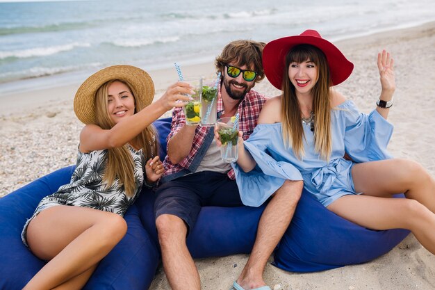 Young hipster company of friends on vacation at beach cafe, drinking mojito cocktail, happy positive, summer style, smiling happy, two women and man having fun together, talking, flirt, romance, three