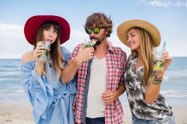 Young hipster company of friends on vacation at beach, drinking mojito cocktail, happy positive, summer style, smiling happy, two women and man having fun together, talking, flirt, romance, three