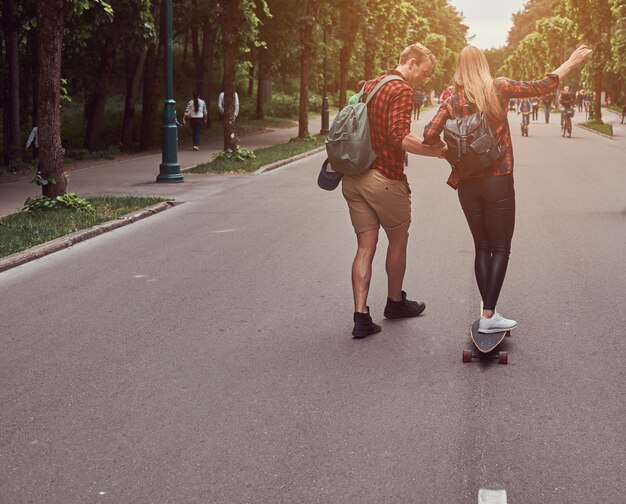 Foto gratuita coppia giovane hipster. guy insegna alla sua ragazza a fare skateboard per strada nel parco.