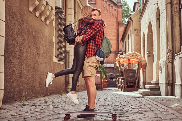 젊은 힙스터 커플, 잘생긴 스케이팅 선수, 그의 여자친구가 유럽의 오래된 좁은 거리에 서서 껴안고 있습니다.