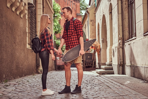 若い流行に敏感なカップル、ハンサムなスケーターと彼のガールフレンドはヨーロッパの古い狭い通りに立っています。