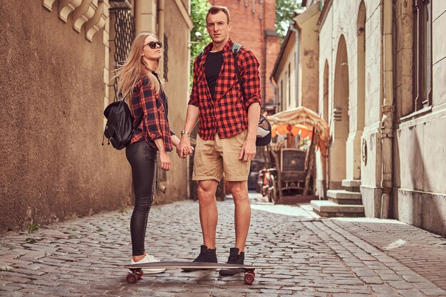 젊은 힙스터 커플, 잘생긴 스케이팅 선수, 그의 여자친구는 유럽의 오래된 좁은 거리에 서 있습니다.