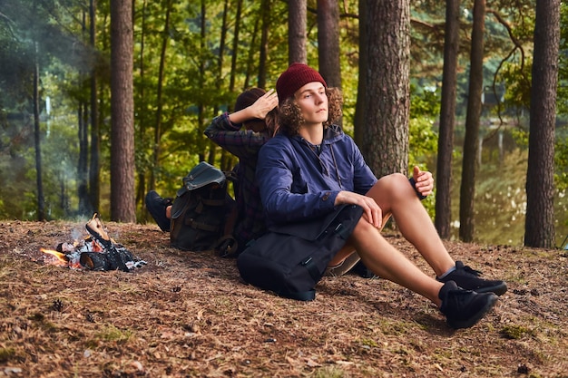 Молодая пара хипстеров путешествует по лесу и сидит спиной к спине, греясь у костра в лагере. Концепция путешествий, туризма и походов.