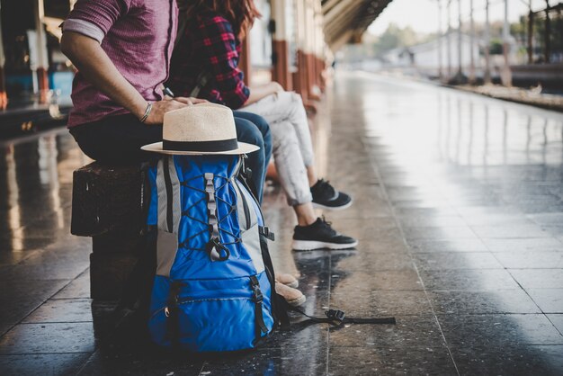列車駅で木製ベンチに座って若いhipsterカップル。プラットフォームで列車を待っているカップル。