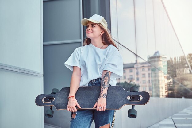 Young hipster girl in cap dressed in white shirt and ripped jeans holds skateboard while posing near skyscraper.