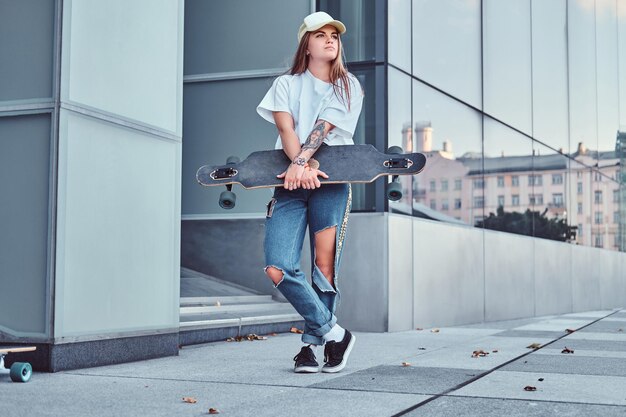 흰색 셔츠와 찢어진 청바지를 입은 모자를 쓴 젊은 힙스터 소녀는 마천루 근처에서 포즈를 취하면서 스케이트보드를 들고 있습니다.