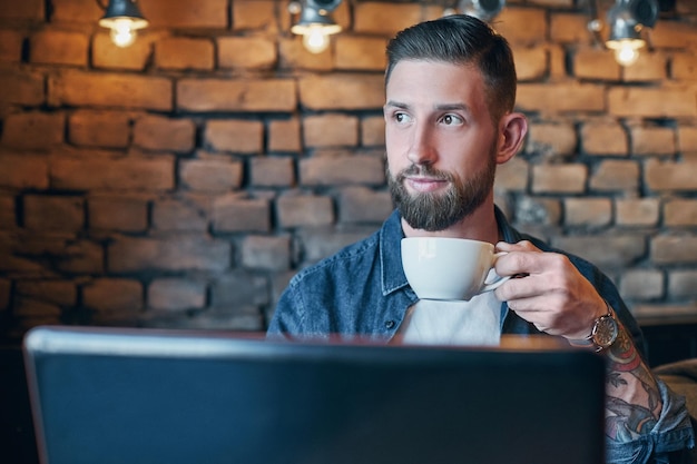 Young hipster guy at the bar having a cappuccino. Young man drinking coffee in city cafe during lunch time and working on laptop